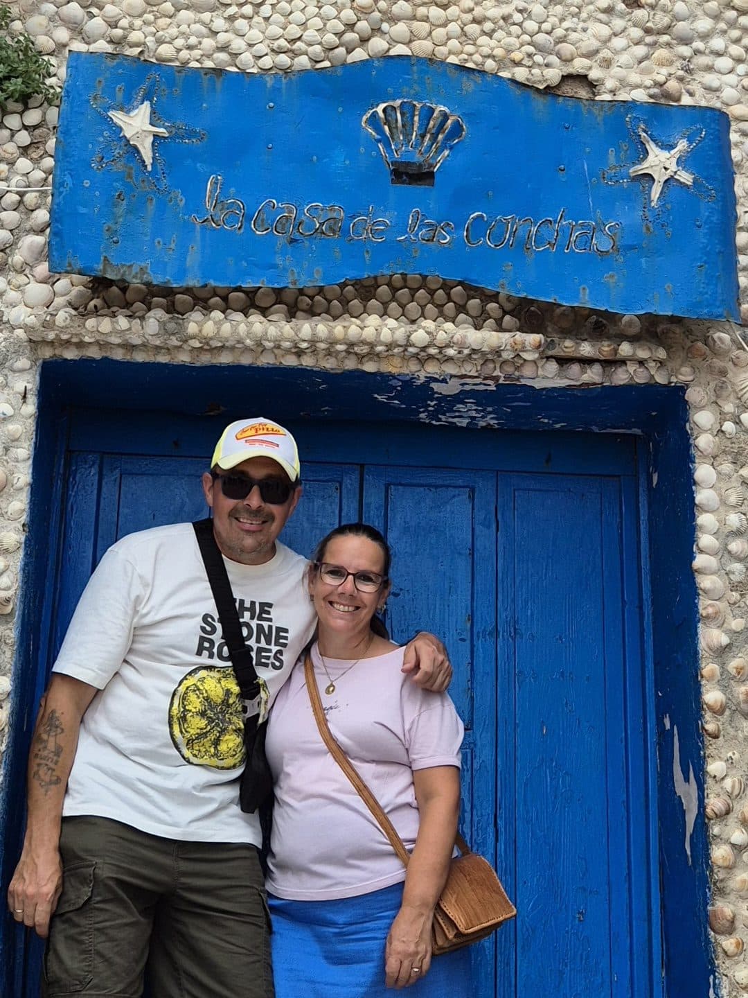 Shane and Yolanda, the founders of Costa Bridge, smiling in front of the blue door of La Casa de las Conchas, a house covered in seashells